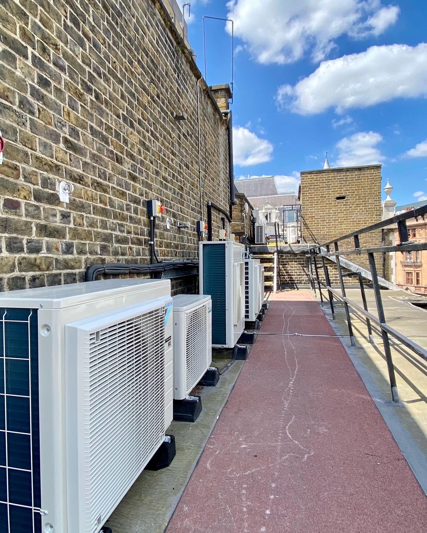 Air conditioning condenser units installed along a rooftop walkway beside a brick building under a blue sky.