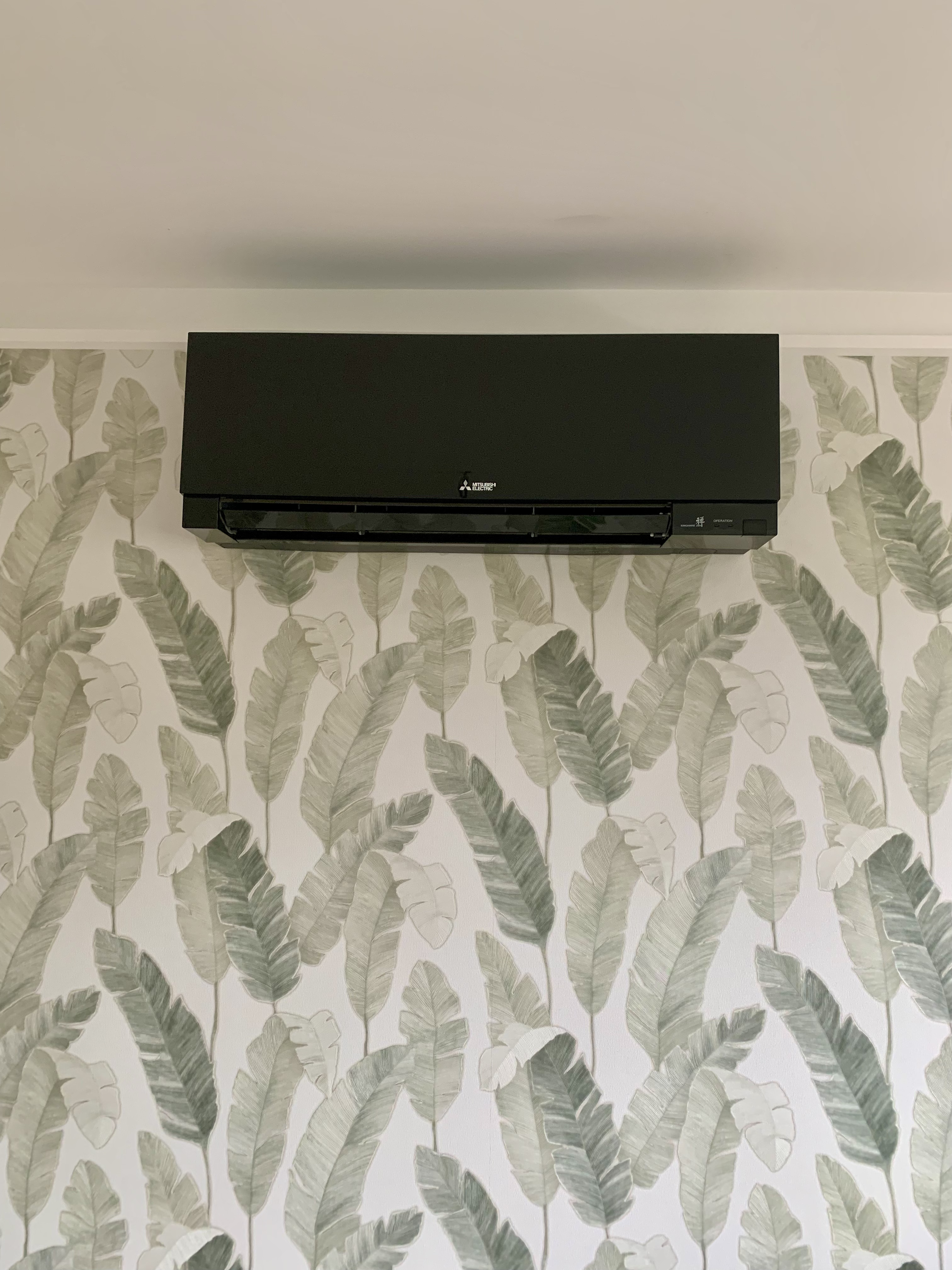 A sleek black Mitsubishi Electric indoor air conditioning unit mounted on a palm leaf patterned wallpaper.