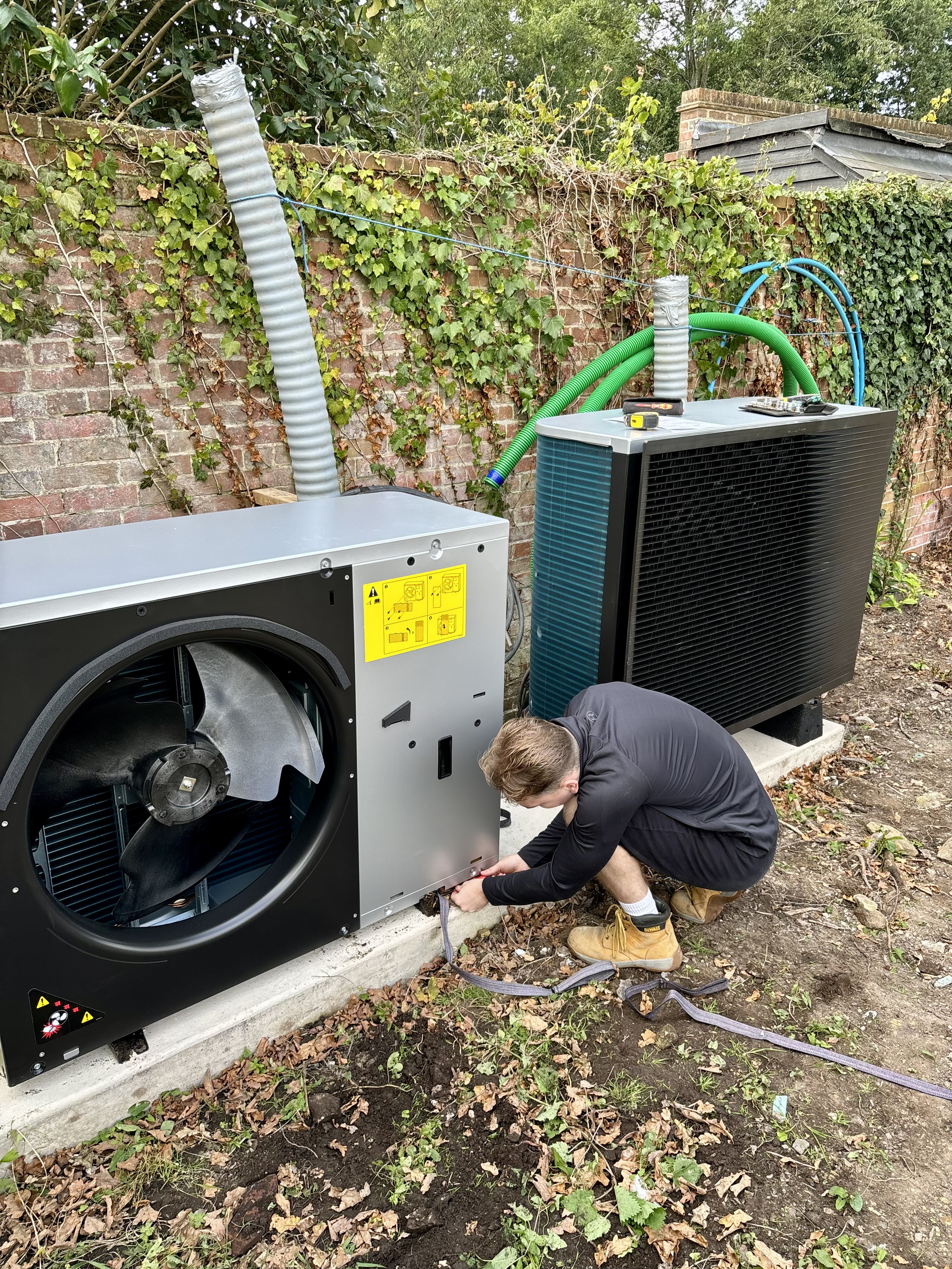 Technician installing an outdoor air source heat pump unit on a concrete base against a brick wall.