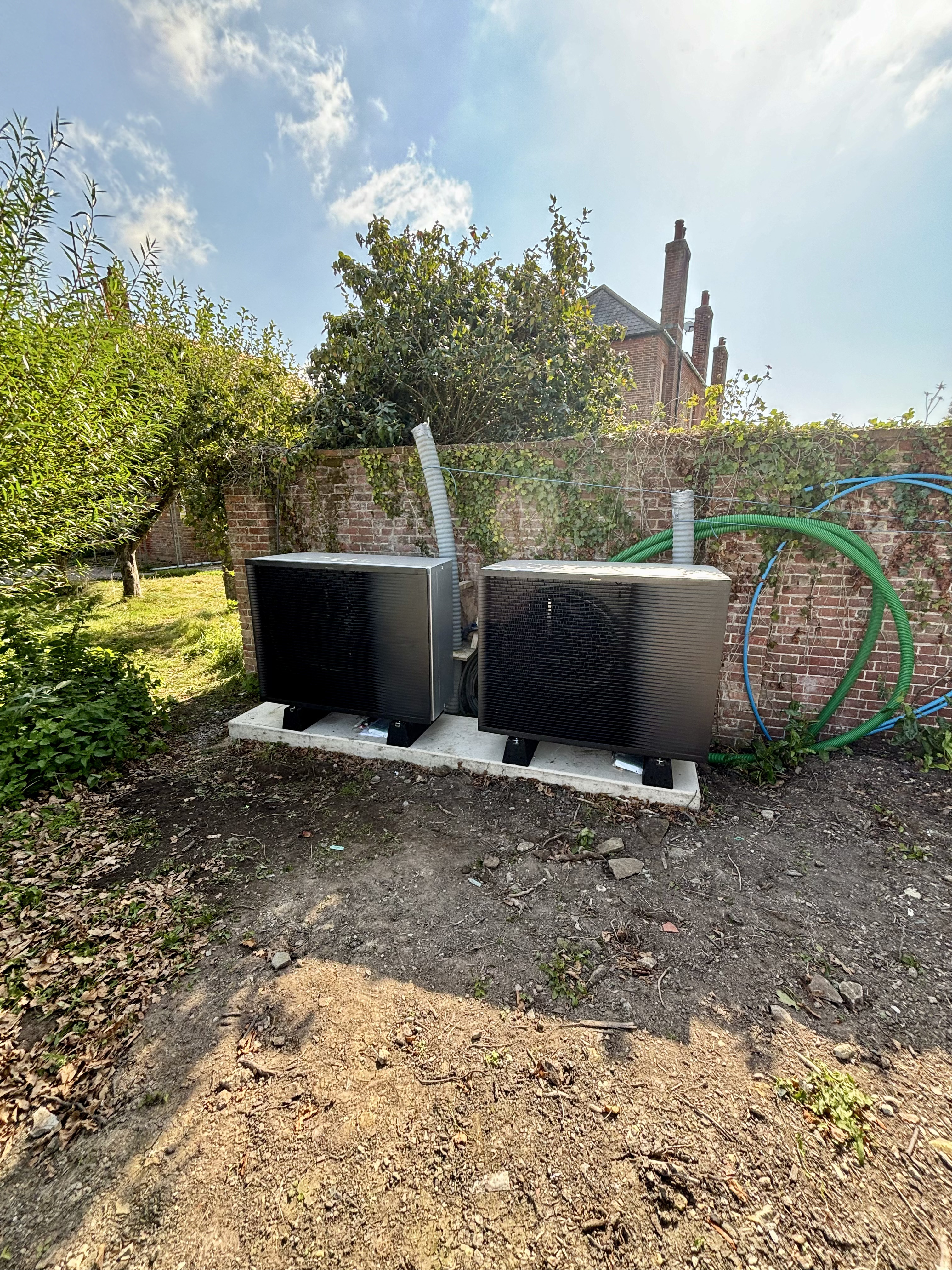 Two outdoor heat pump units installed on a concrete base against a brick garden wall.